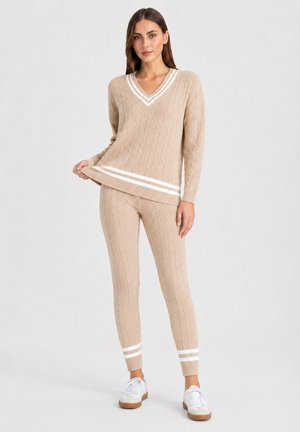 Donna che indossa un maglione beige lavorato a treccia con scollo a V e pantaloni abbinati con strisce bianche, abbinati a scarpe da ginnastica bianche su sfondo semplice.