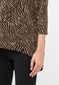 Fekete és arany zebra mintás felső, texturált anyagból, háromnegyedes ujjakkal, fekete, testre simuló nadrággal párosítva.