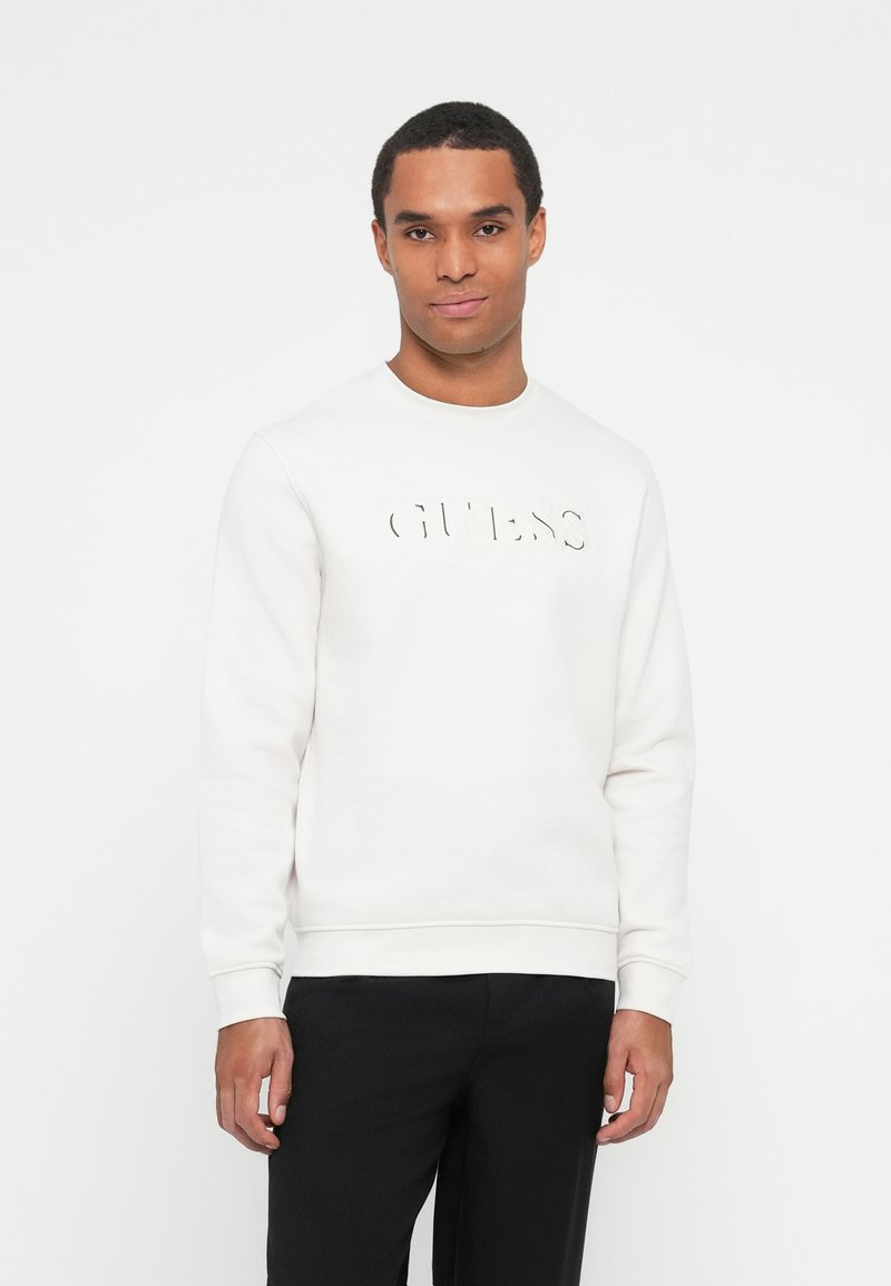 Hombre vistiendo un sudadera blanca con el logotipo "GUESS" en el frente, combinado con pantalones negros, de pie contra un fondo blanco.
