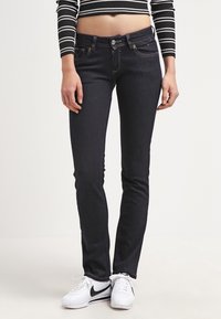 Jeans ajustados de denim azul oscuro, con una cintura media, costuras sutiles y diseño de cinco bolsillos, combinados con zapatillas blancas.