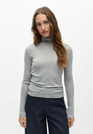 Femme aux longs cheveux bruns portant un pull à col roulé gris clair et un jean bleu foncé, debout devant un fond blanc uni.
