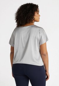 Blusa di raso grigio con maniche corte, scollatura rotonda e una vestibilità morbida, abbinata a pantaloni blu scuro. Tessuto liscio, dettagli di cucitura minimi.