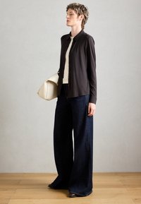 Mørk skjorte med knapper, cremet tanktop og brede mørkeblå denimbukser. En beige håndtaske hviler på armen. Neutral baggrund med trægulv.