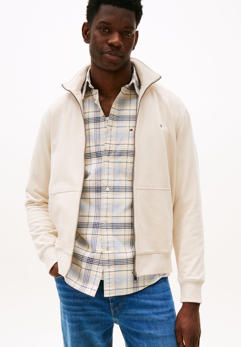 Beige Reißverschlussjacke mit hohem Kragen, über einem hellen karierten Hemd mit Knöpfen in Blau- und Beigetönen. Blaue Denim-Jeans, lässige Passform.