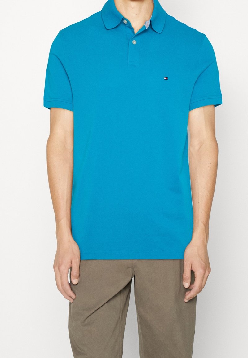 Polo turquoise en coton, doté d'un col classique, de manches courtes et d'un petit logo sur la poitrine. Porté avec un pantalon beige.