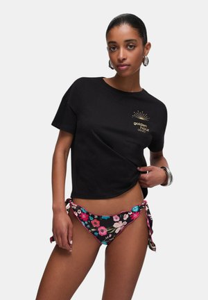 Frau trägt ein schwarzes T-Shirt mit dem Schriftzug "golden hour" und einer Sonnen-Grafik, kombiniert mit schwarzen, geblümten Bikinihosen, die an der Hüfte gebunden sind.