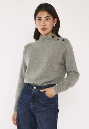 Femme portant un pull en maille gris clair avec des boutons sur l'épaule et un jean taille haute bleu foncé, debout avec une main dans la poche.