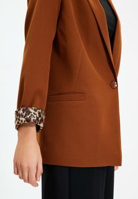 Blazer marrón con un solo botón, con un puño de estampado de leopardo. Diseño simple, corte entallado y un detalle de bolsillo lateral.