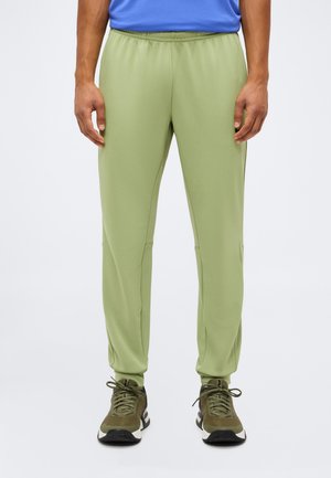 Homme portant un pantalon vert clair coupe décontractée et des chaussures de sport vertes, debout sur un fond uni.