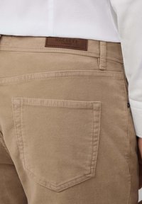 Beige katoenen broek met een gladde textuur, voorzien van een achterzak, leren merklabel en contrasterende stikseldetails.
