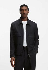 Svart zip-up jacka med krage, med två fickor framtill, gjord av slät tyg, bärs över en vit T-shirt, kombinerad med mörka byxor.