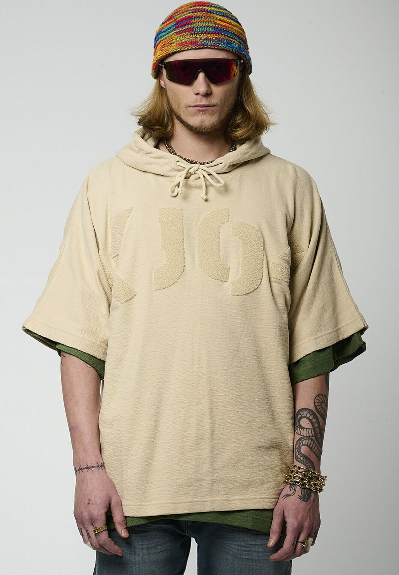 Hoodie oversized beige avec un logo texturé sur le devant, capuche avec cordon de serrage, manches courtes, superposé à une chemise verte, associé à un bonnet coloré.