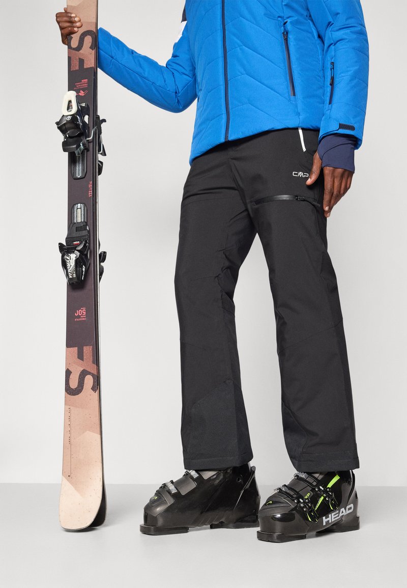 Blauwe geïsoleerde skijas, zwarte waterdichte broek, zwarte skischoenen met groene accenten, houdt een bruine ski vast met zwarte bindingen.