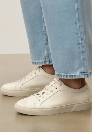 Fehér bőr alacsony szárú fűzős tornacipők világoskék egyenes szárú farmerrel viselve, bézs színű padlón.