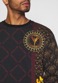 Sort langærmet skjorte med levende guld- og røde mønstre, en cirkulær emblemer og karakteristiske pyntede designs på ærmerne.