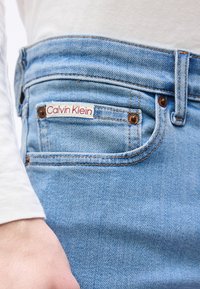 Τσέπη από ανοιχτό μπλε τζιν με λευκή ετικέτα που γράφει "Calvin Klein", φορεμένη με λευκό μακρυμάνικο πουκάμισο που φαίνεται στον καρπό.