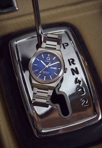 Ceas din oțel inoxidabil argintiu cu quartz, având un cadran albastru, marcaje orare argintii și afișaj pentru calendar, plasat pe un schimbător de viteze metalic.