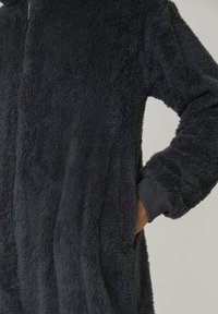 Veste zippée en peluche gris fonc�é avec des poignets côtelés, présentée avec une main partiellement dans la poche latérale sur un fond neutre.