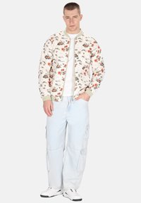 Bloemmotief bomberjack in crème met groene geribde manchetten en kraag, gecombineerd met lichte denim cargopantalon en witte sneakers.