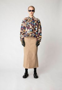 Kleurrijke geprinte jas met verschillende grafische ontwerpen, beige rok, zwarte platform schoenen en zwarte handschoenen tegen een neutrale achtergrond.