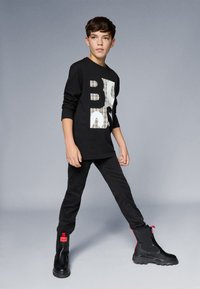 Camisa preta de manga longa com design gráfico branco, combinada com calças pretas e botas de tornozelo chunky pretas com detalhes em vermelho.