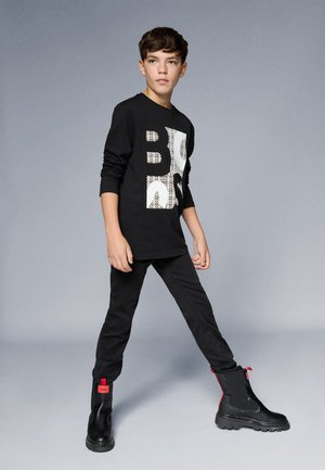 Juodas ilgas rankovės marškinėlis su baltu grafikų dizainu, derinamas su juodomis kelnėmis ir masyviais juodais auliniais batais su raudonais akcentais.