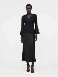 Γυναίκα όρθια με μαύρο κορμάκι δαντέλα μακριάς μανίκι, μαύρη σατέν μίντι φούστα και μαύρα τακούνια με λουράκι αστραγάλου σε λευκό φόντο.