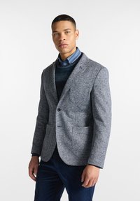 Blazer a motivo houndstooth grigio con rever a punta, due tasche frontali e chiusura a un bottone, indossato sopra un maglione blu e una camicia a righe.