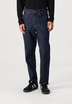 Mørkeblå denimjeans med klassisk rett snitt, som har fem lommer, knappelukking og kontrastsøm, båret med svarte sko.