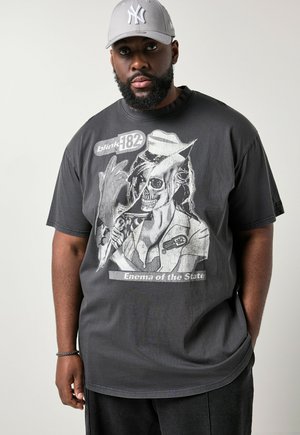 Hombre negro con barba que lleva una gorra gris de los New York Yankees y una camiseta gráfica gris oscuro de Blink-182 "Enema of the State" con un dibujo de una enfermera esqueleto.