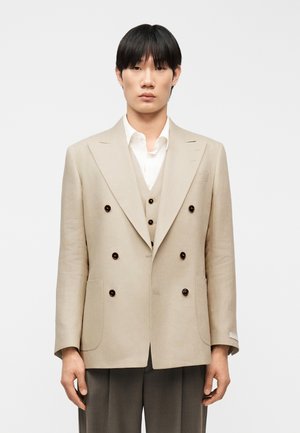 Ung mand iført en beige dobbeltradet blazer og matchende vest over en hvid skjorte, stående mod en ensartet lys baggrund.