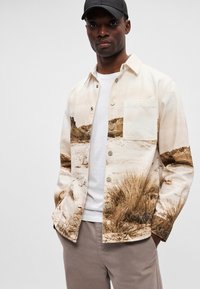 Κοντομάνικο πουκάμισο από ελαφρύ ύφασμα με εκτύπωση τοπίου ερήμου σε μπεζ και καφέ. Διαθέτει εμπρόσθια τσέπη και μεταλλικά κουμπιά.