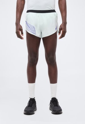 Sportoló fehér futóshortot visel kék mintával és logóval, fehér bokazoknit és fekete futócipőt, egy egyszínű háttéren áll.