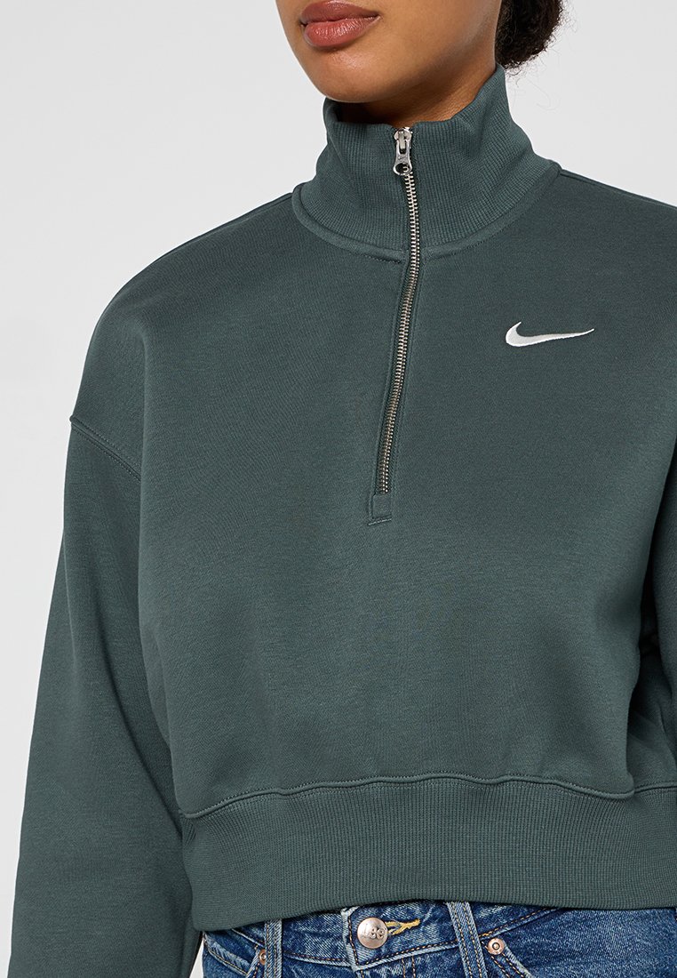 Sudadera recortada de color verde oscuro, con un cuello alto y una cremallera. Cuenta con un pequeño logo blanco de Nike en el lado izquierdo. Textura suave.