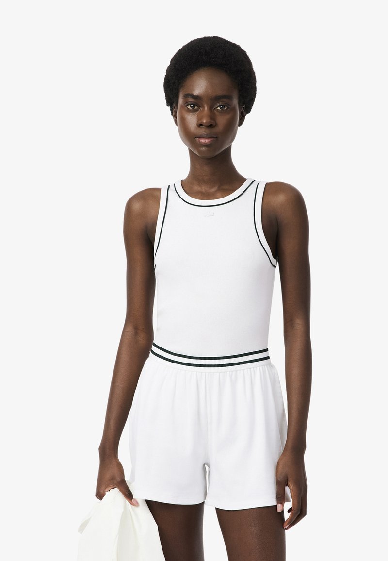 Vrouw met kort krullend haar, gekleed in een witte mouwloze top en witte shorts met zwarte bies, die een witte doek in haar linkerhand vasthoudt.