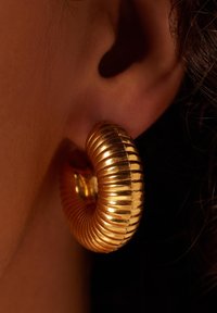 Gouden ringoorbel met ribbeltextuur. Het ronde ontwerp zit nauw tegen het oor, waardoor het glanzende, reflecterende oppervlak wordt benadrukt.