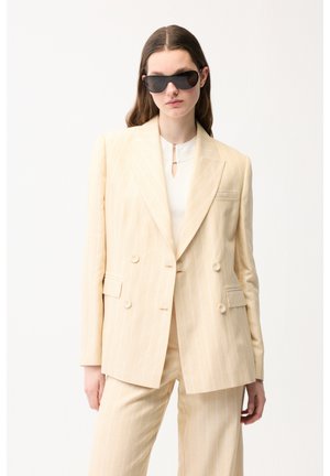 Kvinde iført beige dobbeltradet jakkesæt med striber og matchende bukser samt store sorte solbriller, stående foran en hvid baggrund.