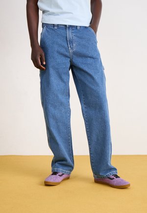 Personne portant un jean bleu ample et des baskets violettes debout sur un sol jaune avec une main dans la poche.