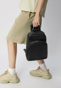 Mochila de cuero negro con dos compartimentos con cremallera, un asa superior y una textura suave, sostenida por una persona que lleva zapatillas beige y una falda.