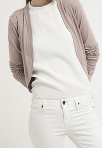 Jasnoróżowy kardigan nałożony na fakturowany biały top, w zestawieniu z białymi dżinsami. Kardigan ma długie rękawy z marszczeniami przy mankietach.