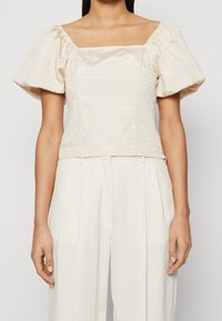 Haut à manches ballon beige clair avec un décolleté carré, en tissu texturé, au design cropped et avec des embellissements minimaux, associé à un pantalon crème à jambes larges.