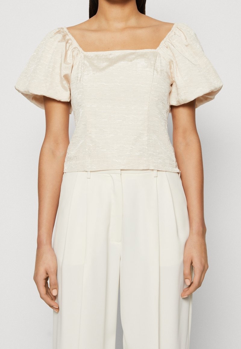 Haut à manches ballon beige clair avec un décolleté carré, en tissu texturé, au design cropped et avec des embellissements minimaux, associé à un pantalon crème à jambes larges.