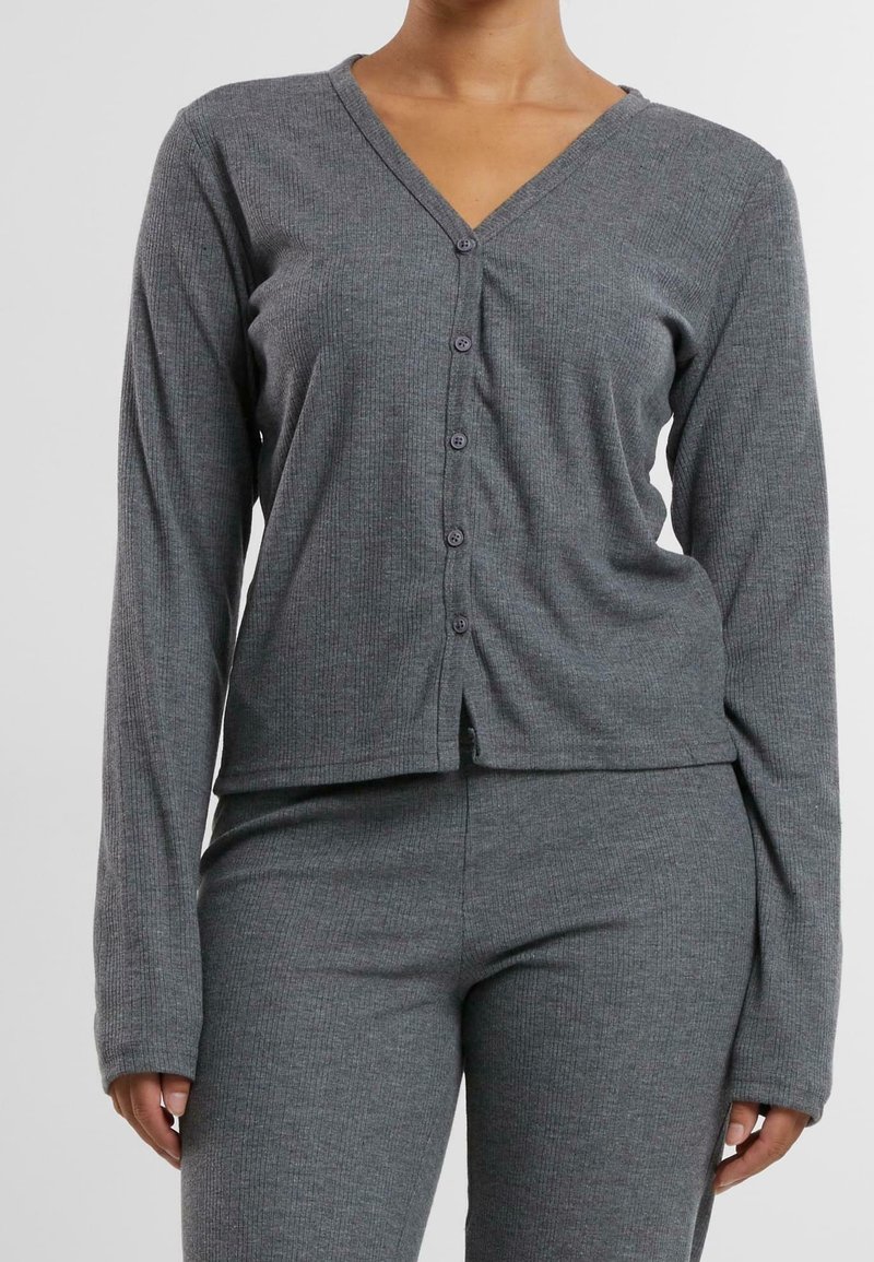 Femme portant un haut côtelé gris à manches longues avec boutons et un pantalon assorti, montrée de la poitrine aux hanches sur un fond uni.