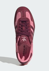 Sneaker aus burgunderrotem und pinkfarbenem Wildleder mit abgerundeter Spitze, ausgestattet mit Stoffschnürsenkeln und drei dunklen Streifen. Logo auf der Zunge sichtbar.