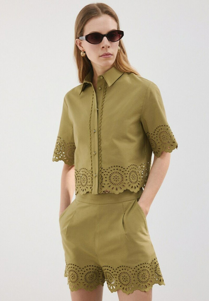 Femme portant une chemise à manches courtes et un short assortis de couleur vert olive avec des détails en dentelle découpée en cercle, des lunettes de soleil noires et des boucles d'oreilles créoles dorées.