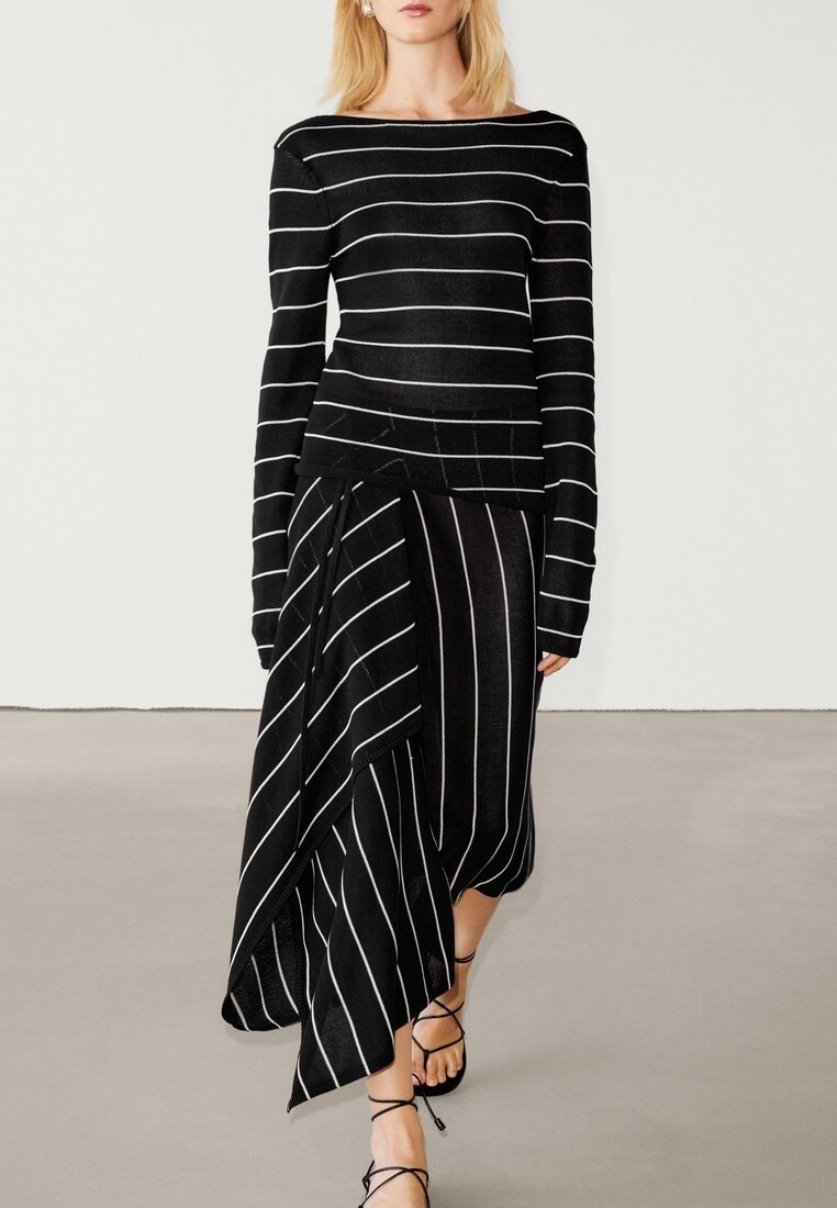 Femme portant un haut noir à manches longues et une jupe asymétrique à rayures blanches, associée à des sandales noires à lacets.