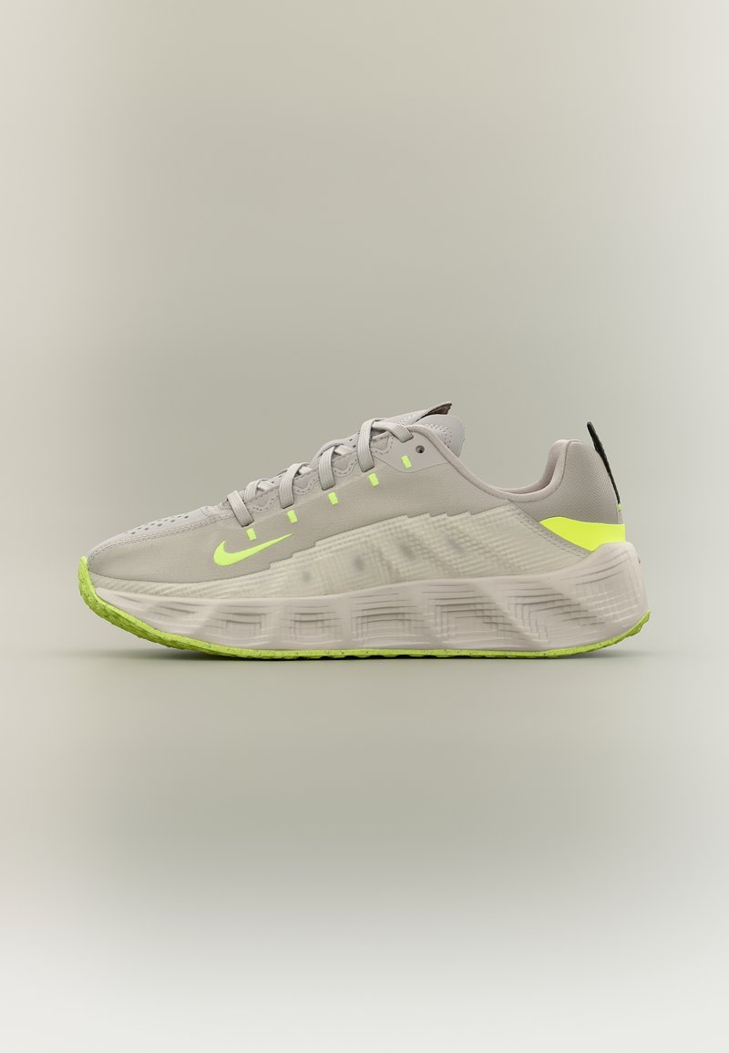 Ανοιχτό γκρι αθλητικό παπούτσι με νεον κίτρινο σήμα Nike, λεπτομέρειες και σόλα, με υφή χονδρής σόλας και κλείσιμο με κορδόνια, πλάγια όψη.