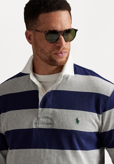 Marineblauw en grijs gestreept rugbyshirt met een witte kraag, voorzien van een in het groen geborduurd logo. Model draagt ronde zonnebril.