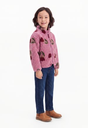 Enfant aux cheveux mi-longs portant une veste polaire rose à fleurs, un jean bleu et des bottines marron, debout et souriant.