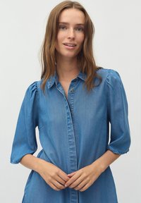 Denimska sraj�čna obleka v svetlo modri barvi z gumbom spredaj, kratkimi puhastimi rokavi in sproščenim krojem. Ima mehko teksturo in stranske žepe.